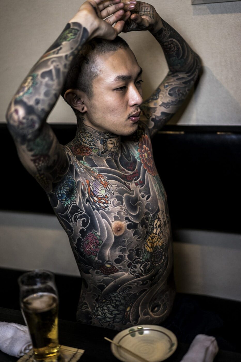 Young Japanese Man Exposing His Tattoos In A Japanese Bar, he is shirtless and has his arms raised above his head, his whole torso is covered in elaborate tattoos
