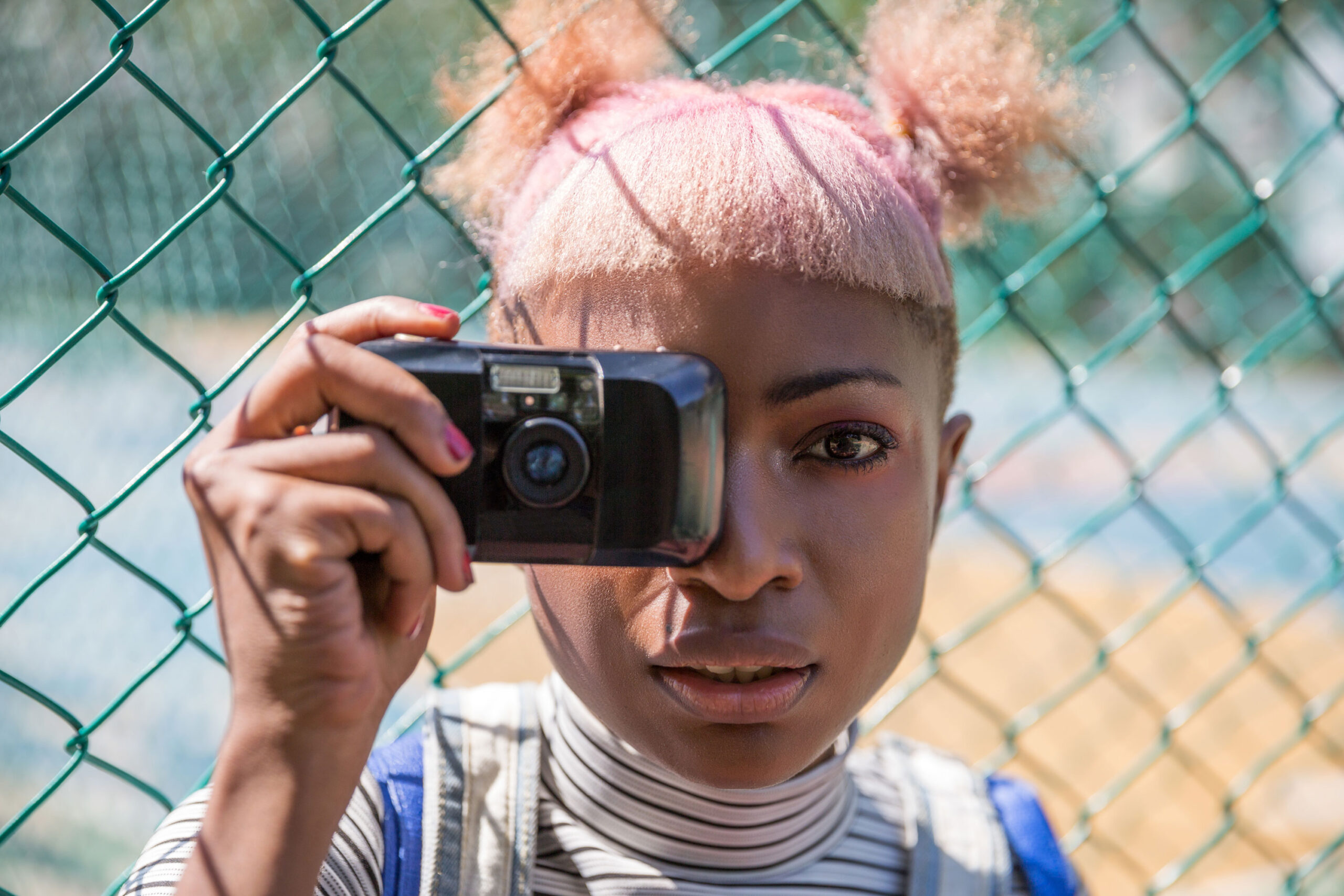 Creative black girl taking photo of viewer with film camera near fence. Her hair is pink and she has a funky haircut