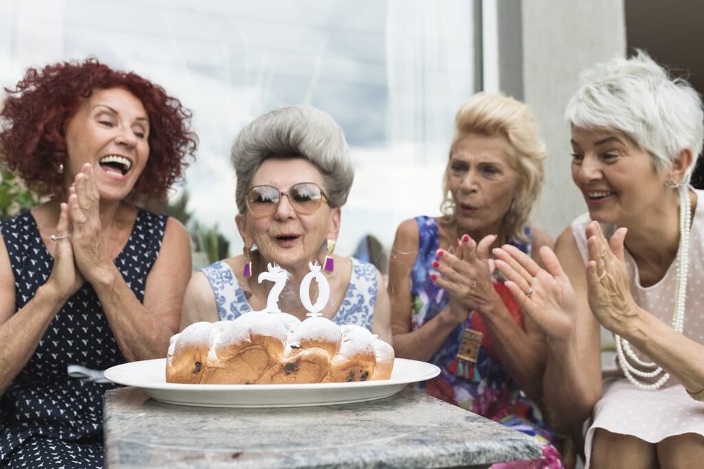 Four older white women clap together around a 70th birthday cake