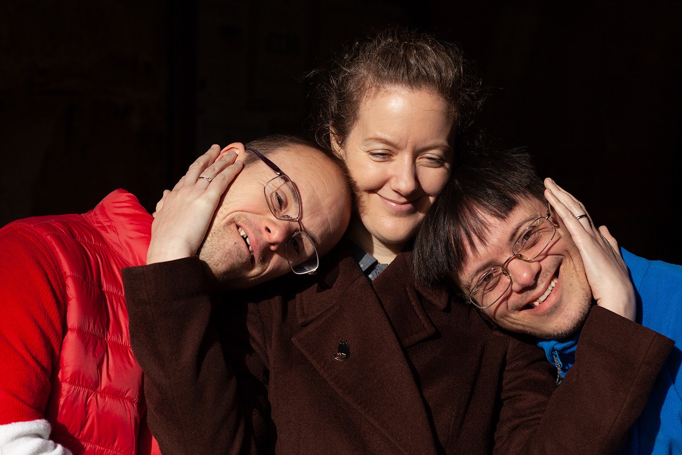 Three adoptive siblings, two with down syndrome share an affectionate happy moment. The sister in the middle puts her arms lovingly around her brother's faces.