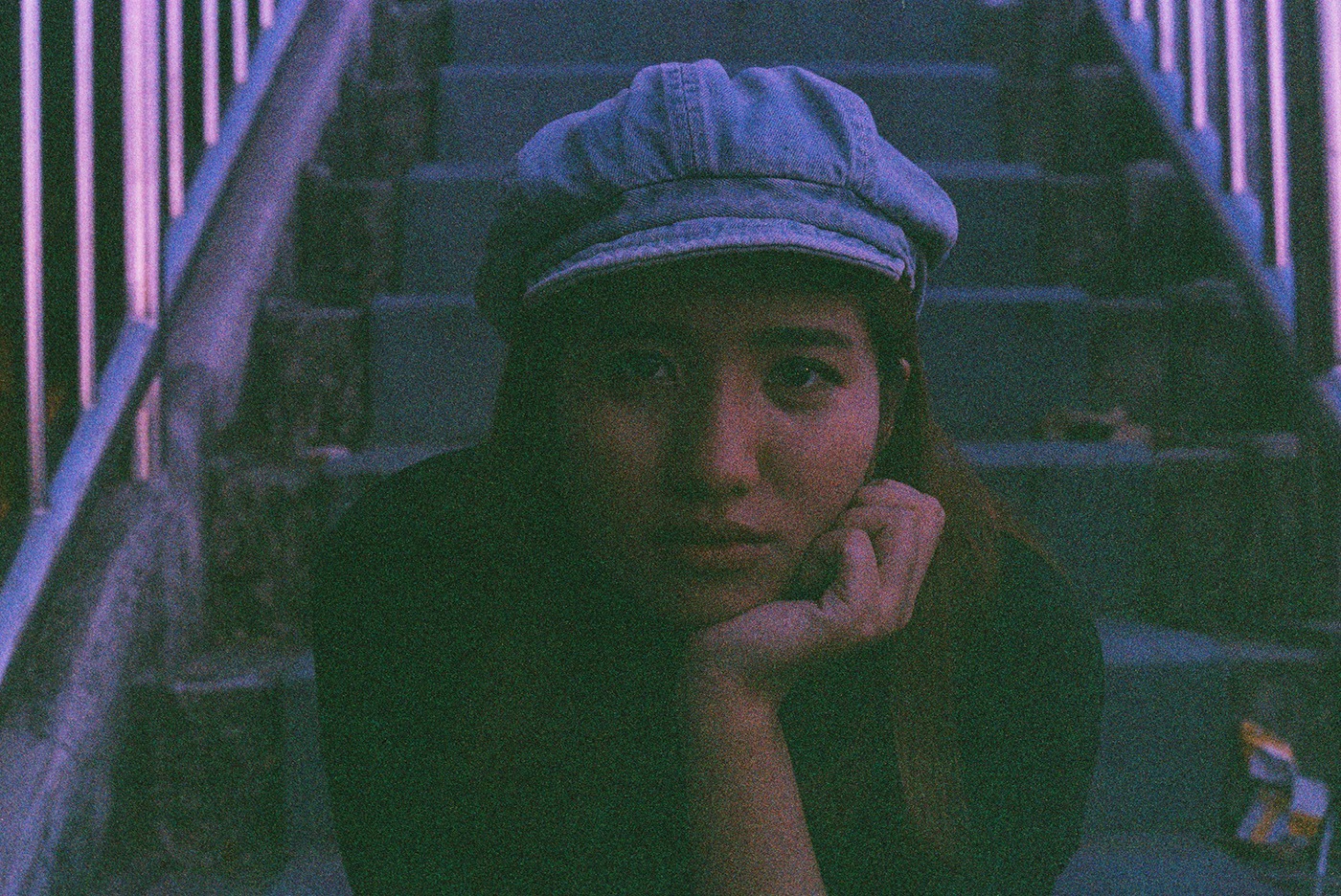 Young Asian woman in an urban setting, sitting on concrete steps staring into the camera. Heavy grain and color shifts from being shot on expired film.