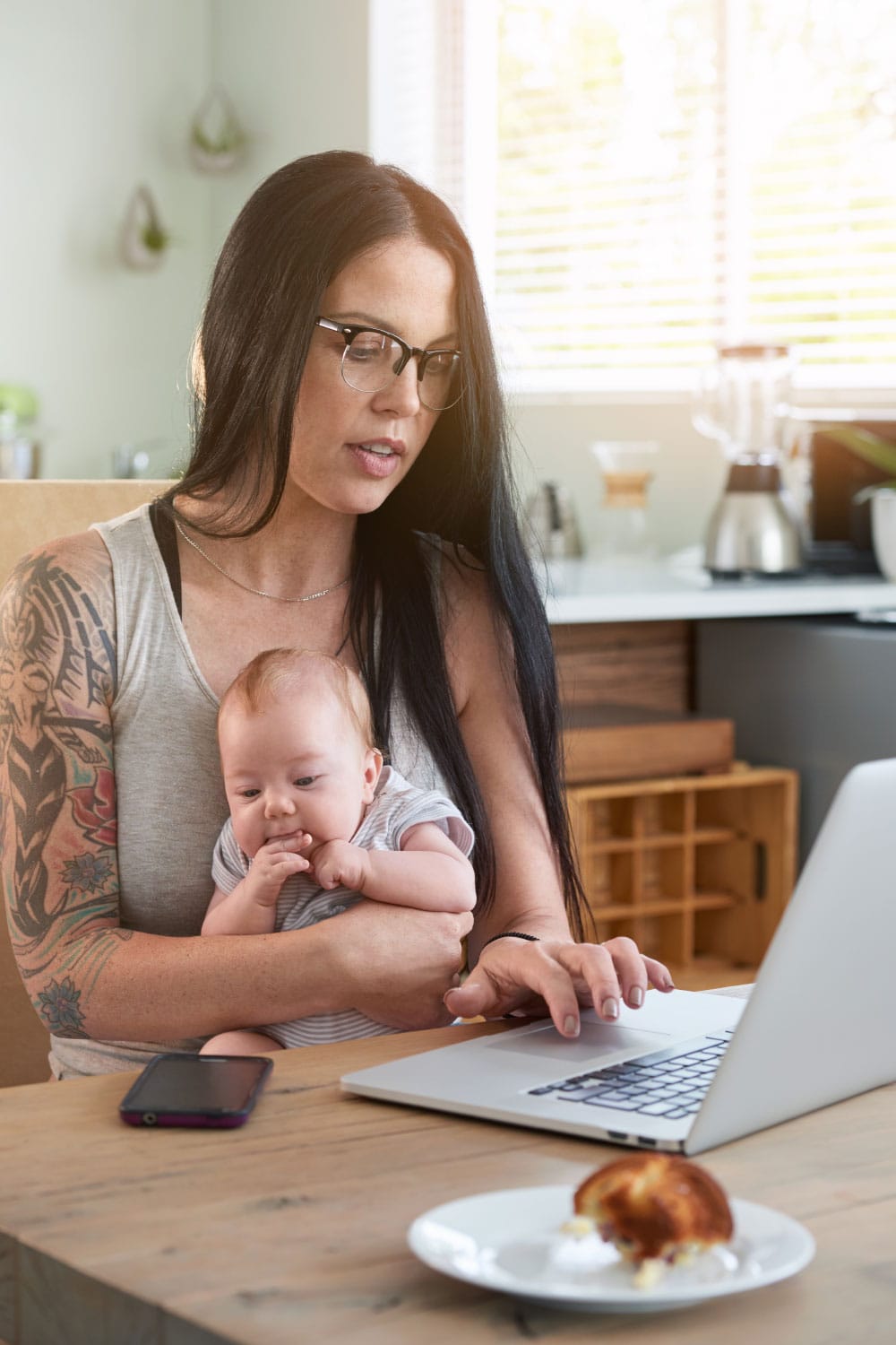 Mother Working From Home With Baby. Work from home mum holding baby while working on laptop computer, busy multitasking during maternity period