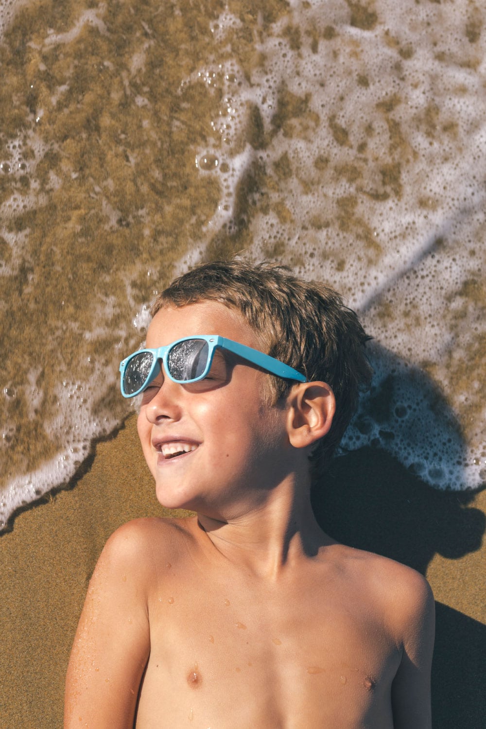 Little boy lying on sand on the beach looking at the sun and smiling