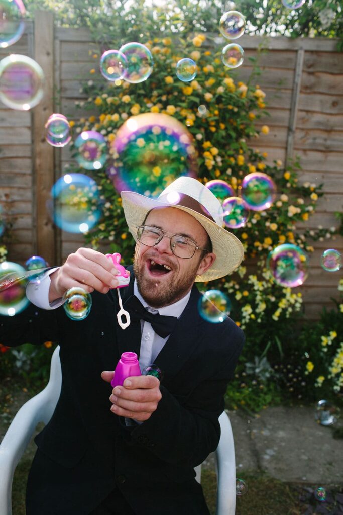 A man with Down's Syndrome in his thirties smartly dressed in a hat and bow tie laughs delightedly while blowing bubbles in a sunny garden.