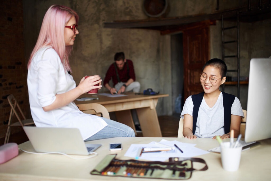 Young white female stylist with pink hair sitting on table and talking to her smiling Asian colleague in loft office