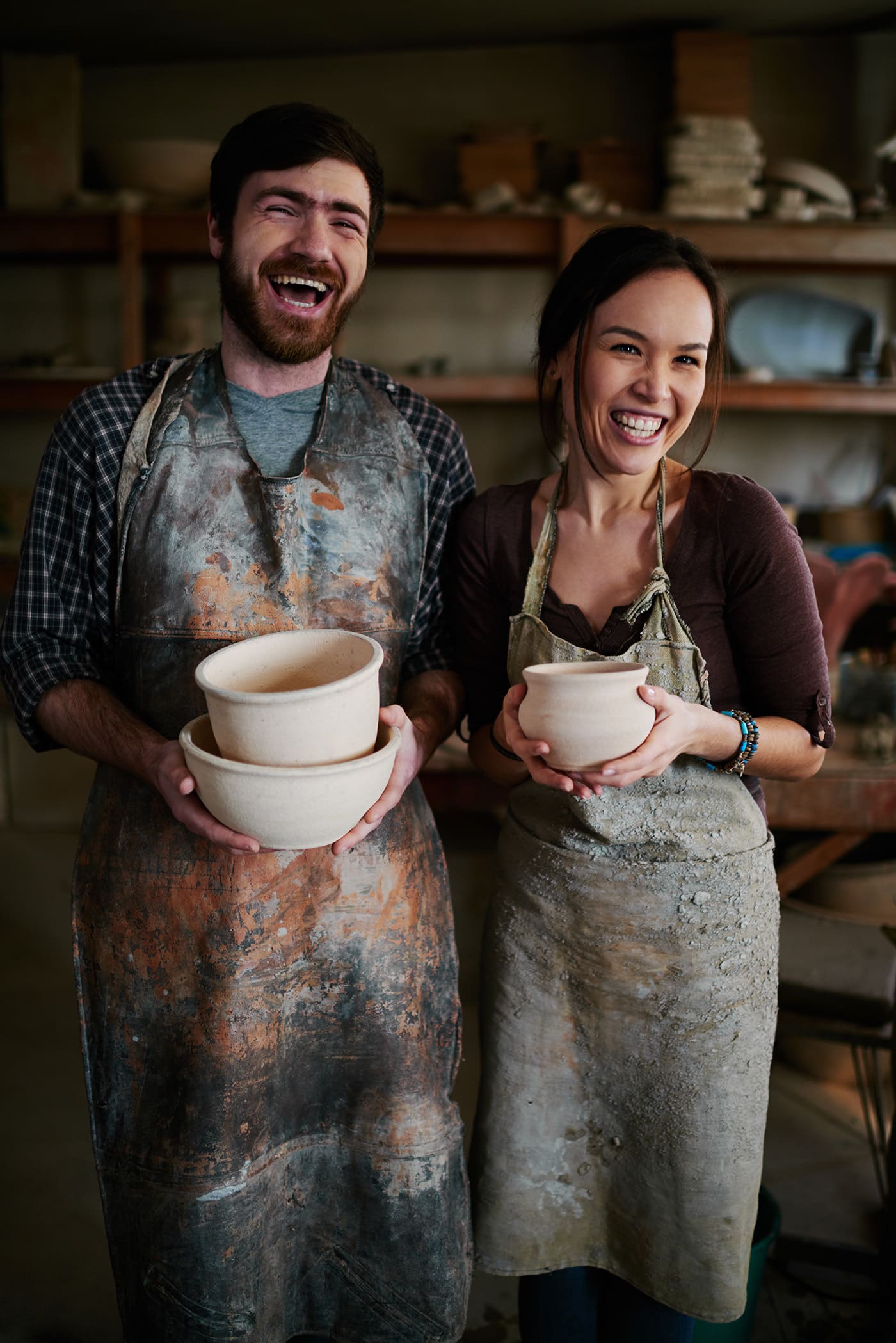 Warm toned portrait of happy potters family posing with clay ceramics in small workshop and laughing genuinely