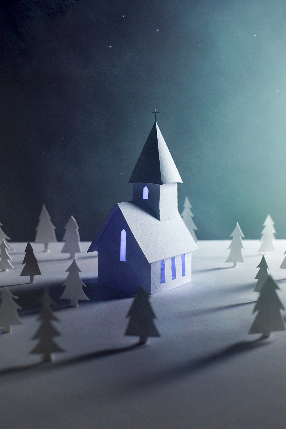 A little paper church with its lights on sits in a wooded area