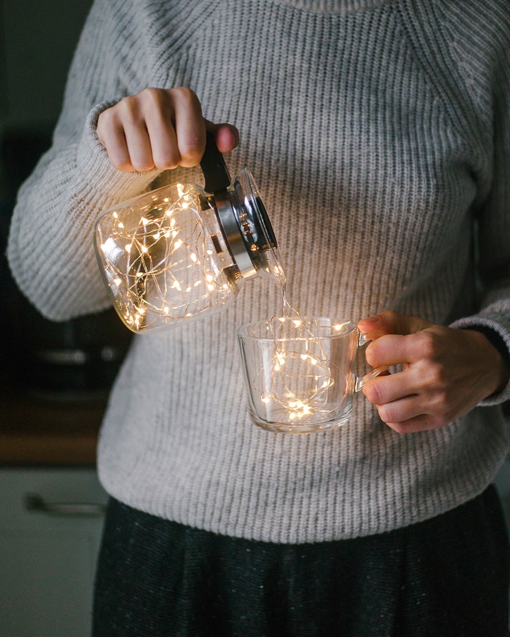 Faceless shot of woman holding glass cup and pouring glowing garland from pitcher.