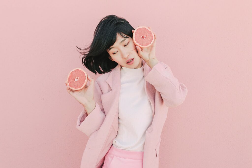 Woman with medium length dark hair wearing all pink, poses with pink grapefruit halves infront of matching pink wall