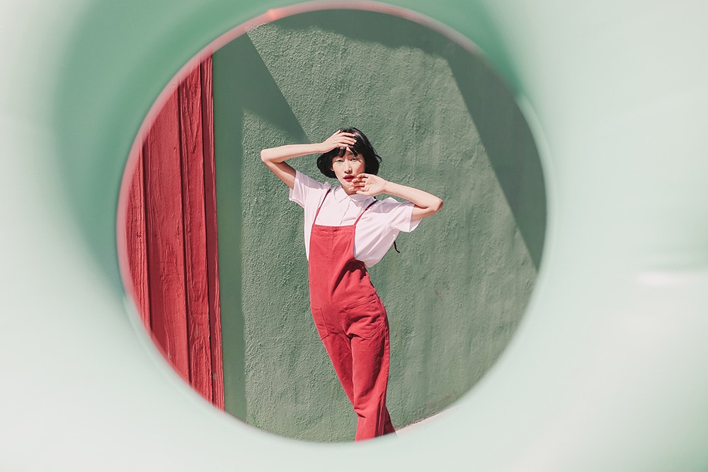 Girl poses in mirror wearing red overalls in front of green stucco wall