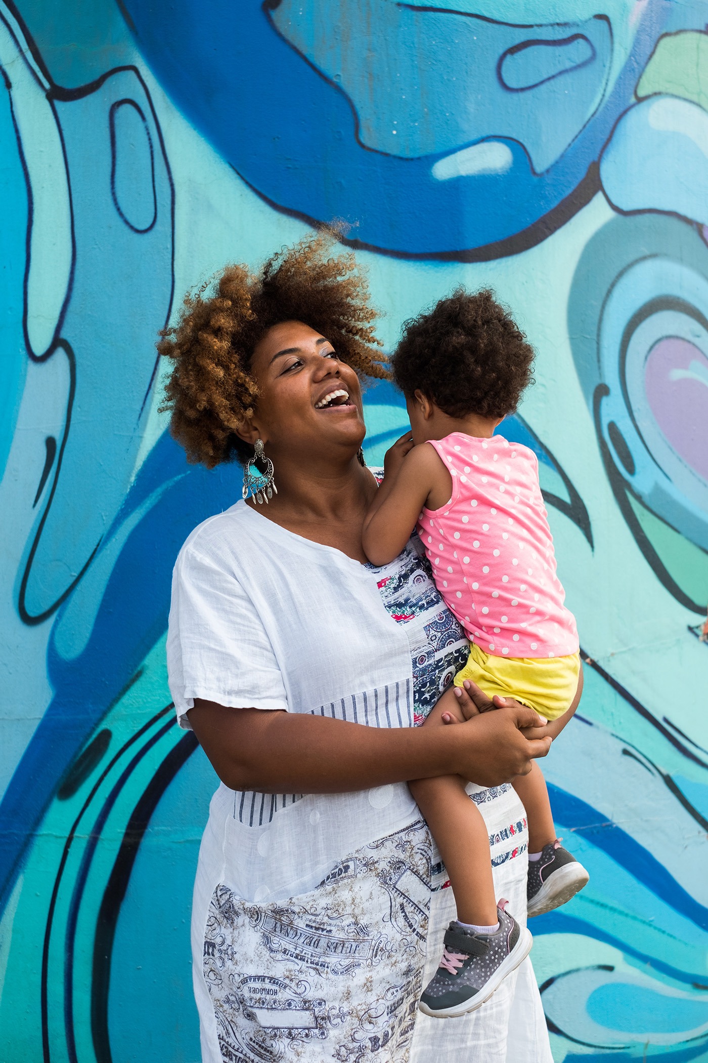 Smiling woman with standing in front of the wall with graffiti and holding an anonymous two-year-old child.