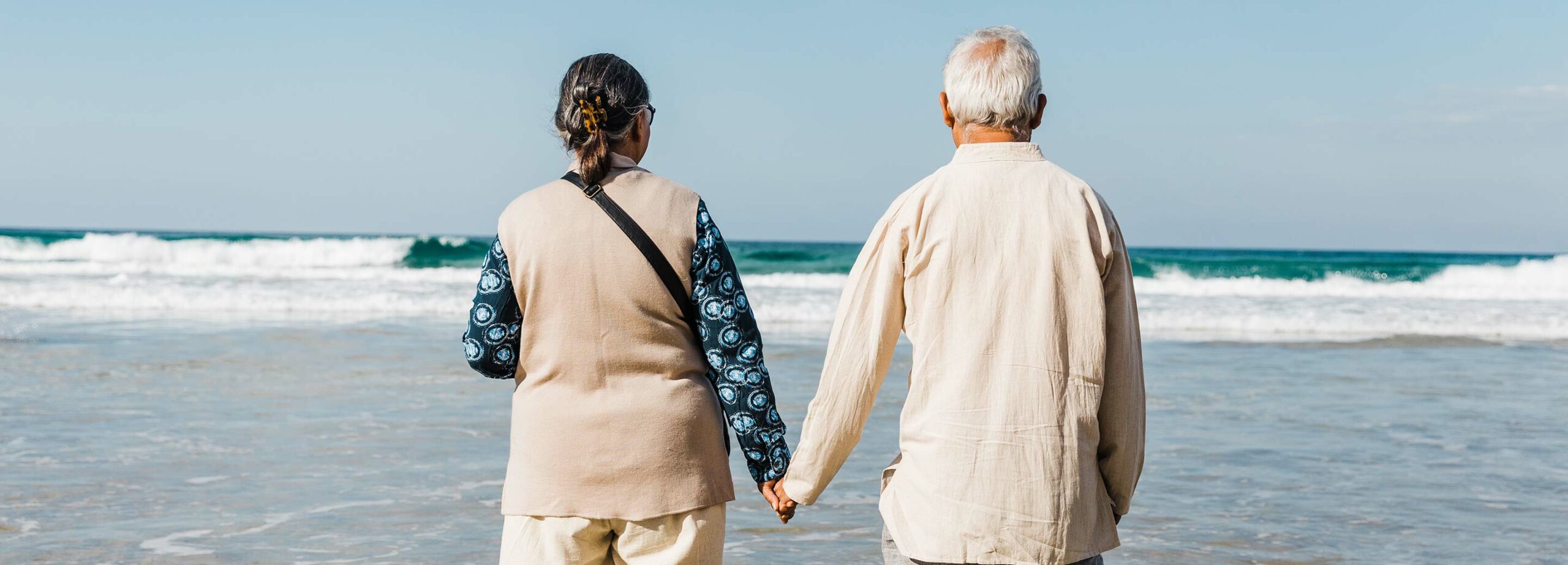 Senior South Asian couple holding hands on a beach facing the ocean. Elderly couple travel around Australia.