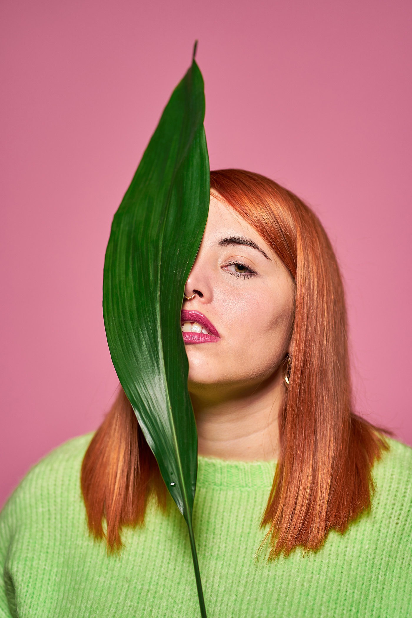 Curve model with red hair keeping green leaf near face and looking at camera against pink background