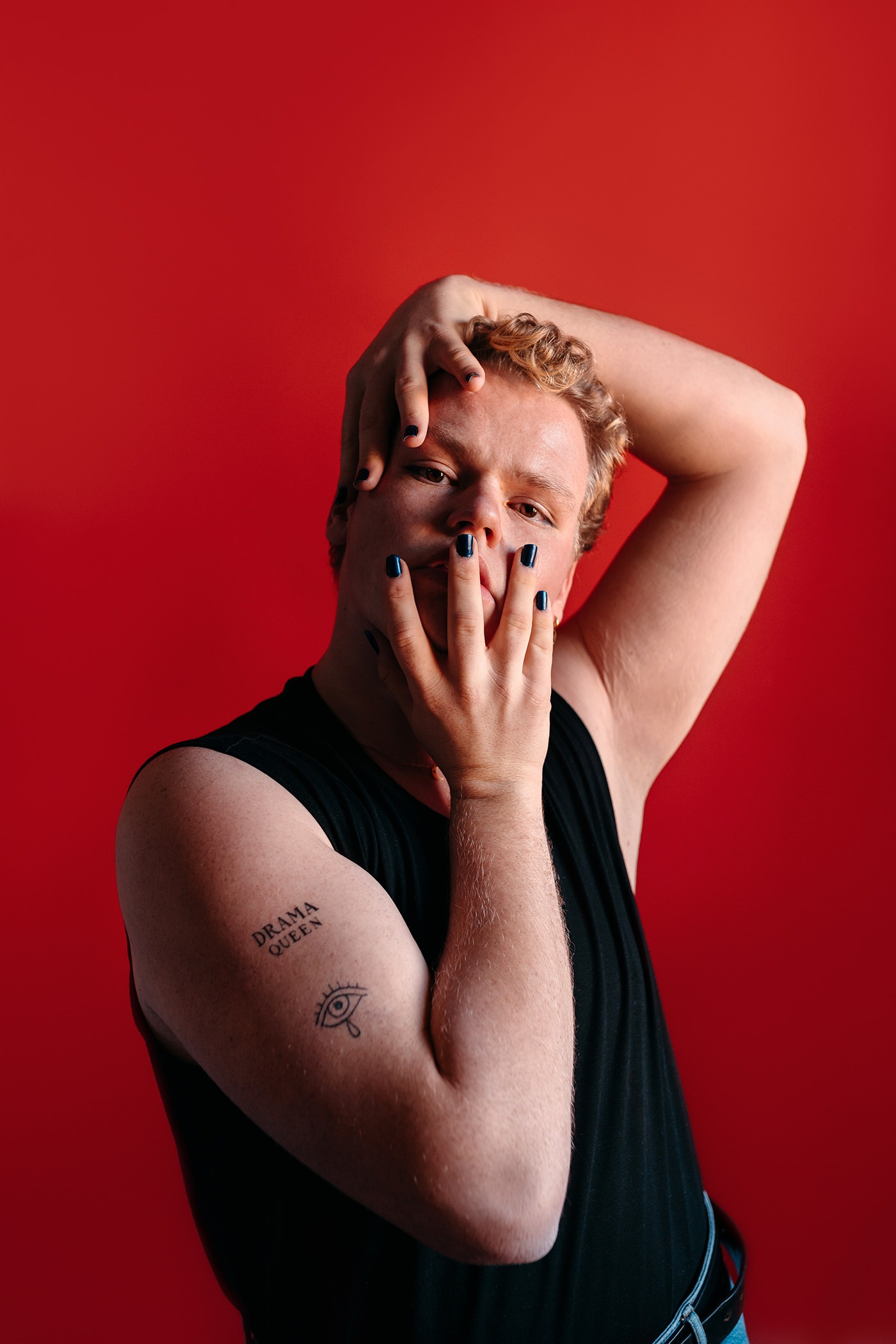 Studio portrait with red background of a young blonde gay man wearing a tank top and dark nails, looking at camera.