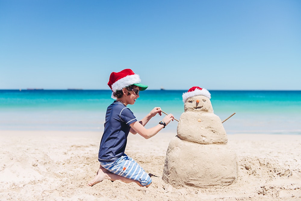 Boy in a bathing suit and Santa hat making a sand snowman at the beach on Christmas day in Australia