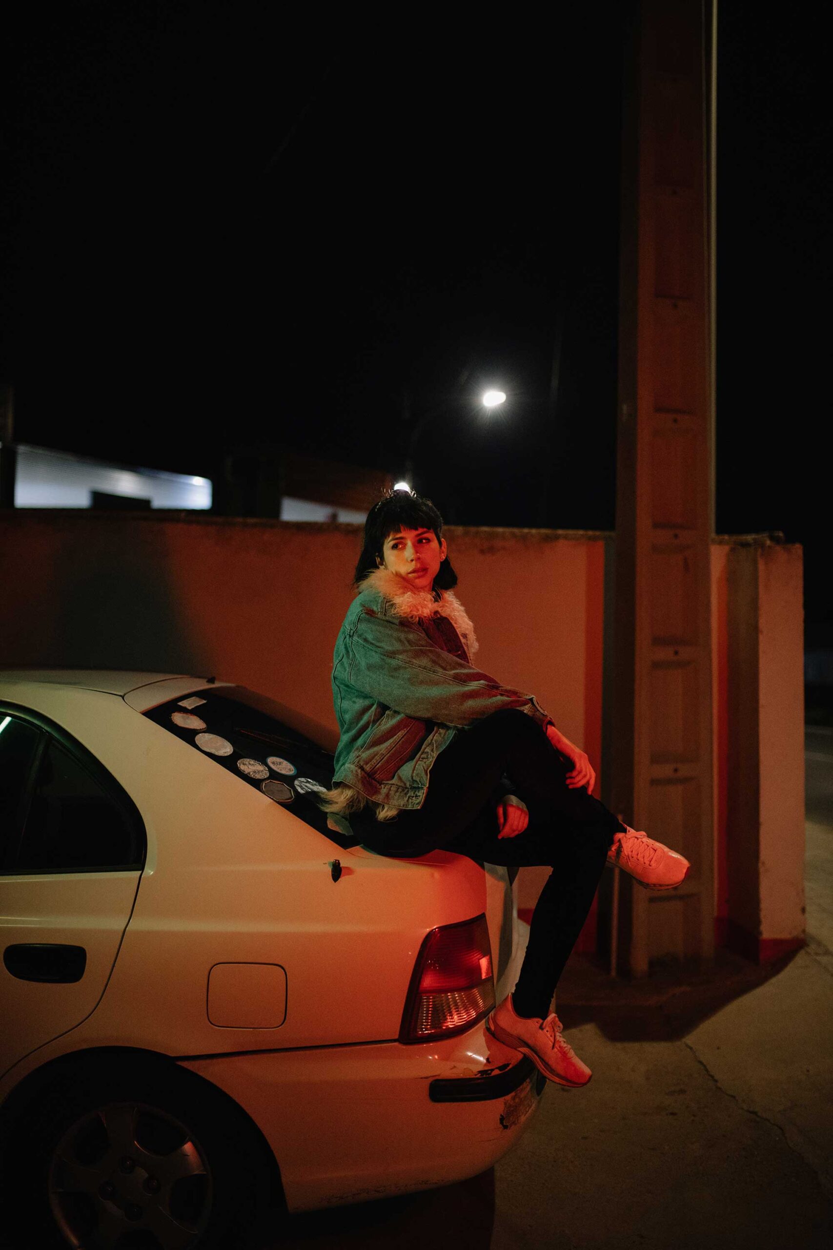 Stylish Woman On Cur Trunk In Nigh Young girl in denim jacket sitting on car trunk in nighttime looking away in neon light.