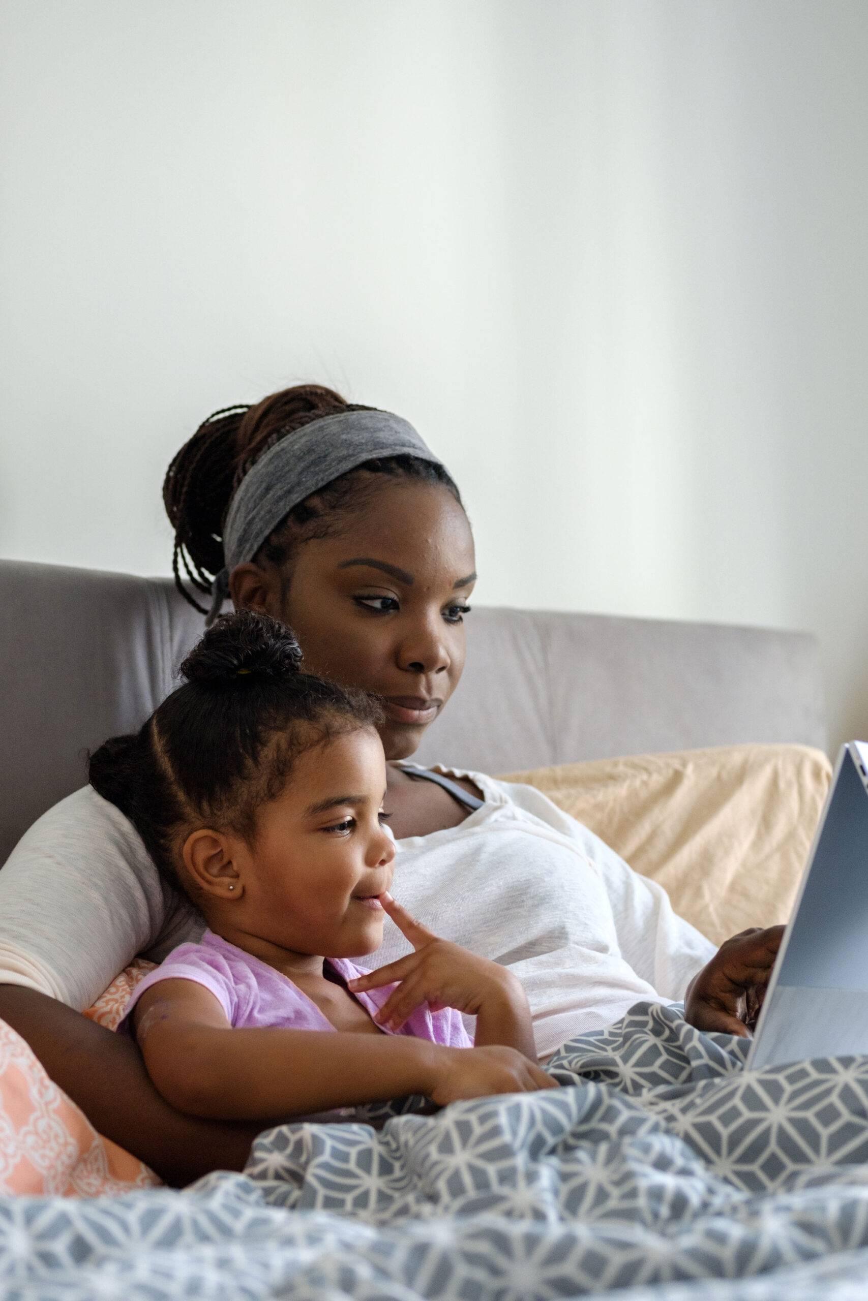 African American mother with her mixed race daughter laying on the parents bed watching educational shows on their 3 in one computer device. This computer is an all in one device that can be used as a laptop, tablet or entertainment device. The lighting in this scene is coming from the window.