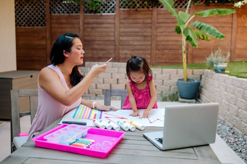 Mid-30's mother with hearing aid helps 2 yr old daughter with art project in beautifully landscaped backyard.