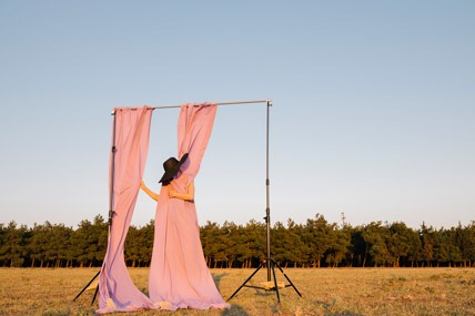 Anonymous woman draped in pink fabric from photography setup wearing large black hat, standing in field at golden hour