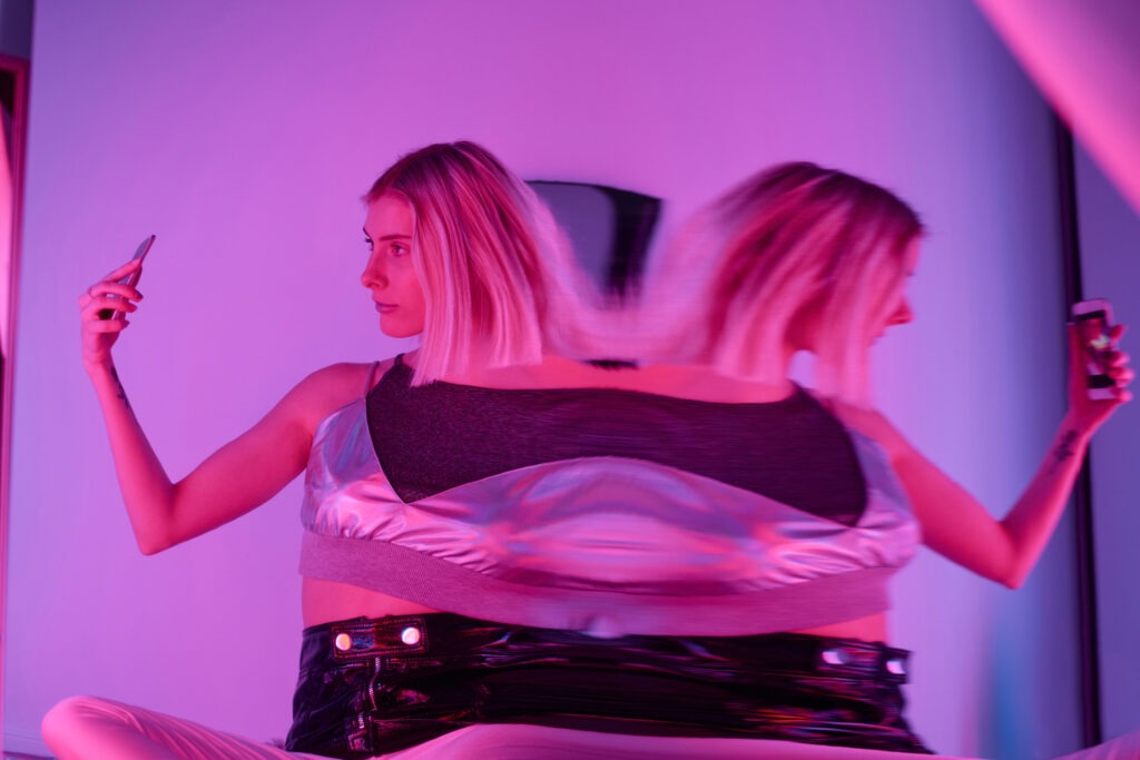 Modern blonde in fractal reflection taking selfie with smartphone sitting in pink neon lights