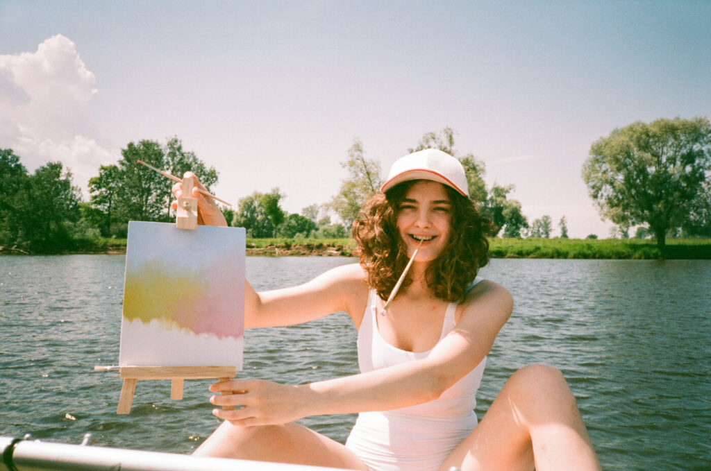 Girl In White Swimsuit With A Pencil In Her Mouth Show Her Artwork In Process