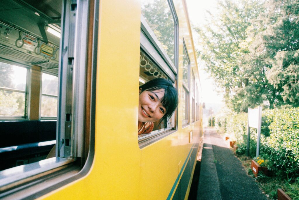 Young woman peering head out the window and smiling at the camera from a bright yellow retro train