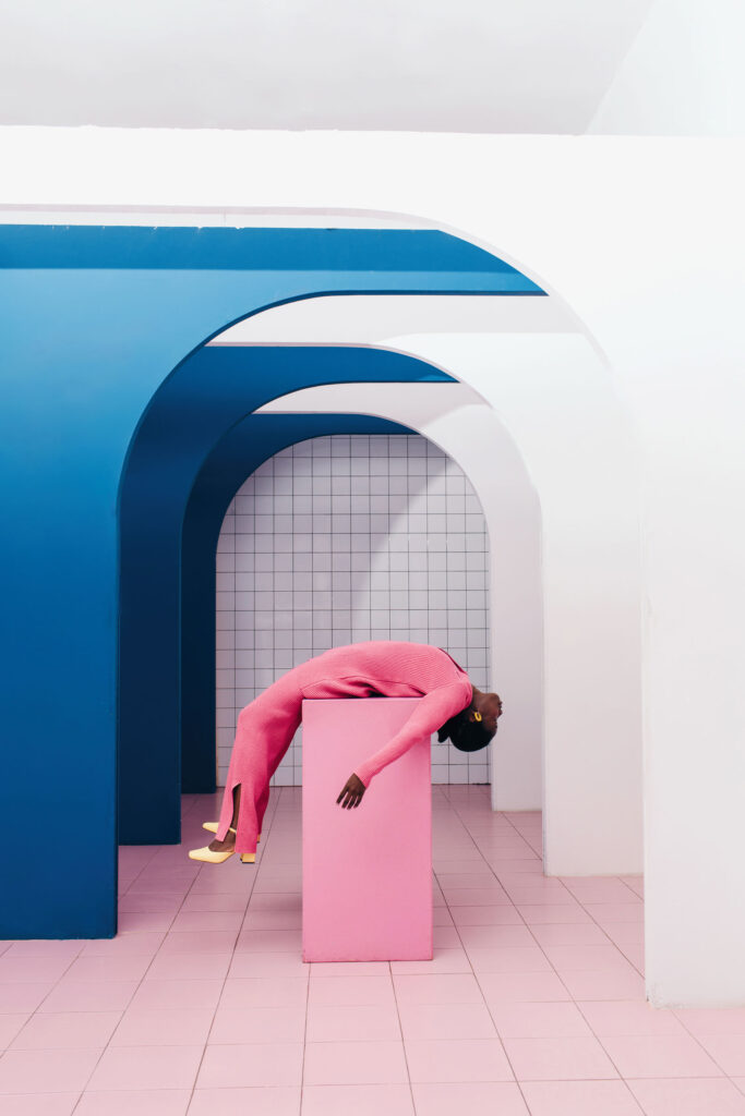 Side view of eccentric black female in stylish pink clothes lying on cube under arched doorway in minimal interior. Research of interaction of body and space.