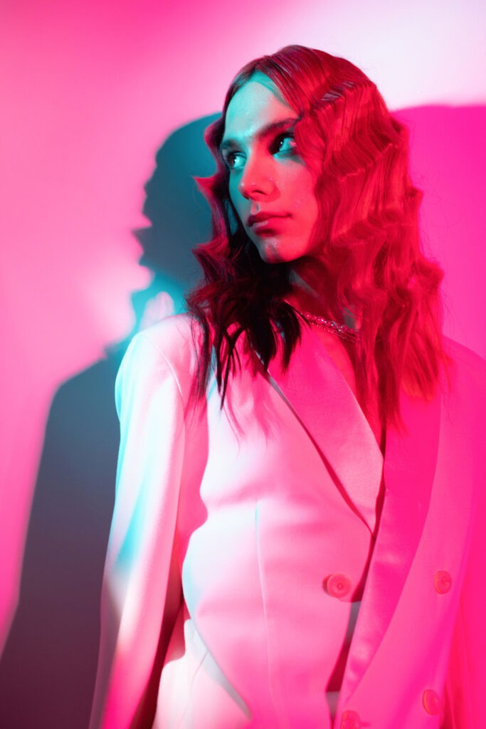 Studio portrait of transgender woman with long wavy hair wearing a white suit in vibrant pink and cyan lights