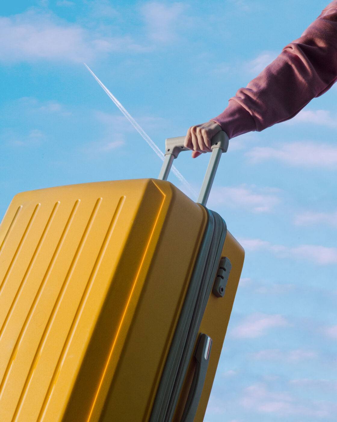 An unrecognizable person holds a travel yellow plastic suitcase by the handle against a blue sky