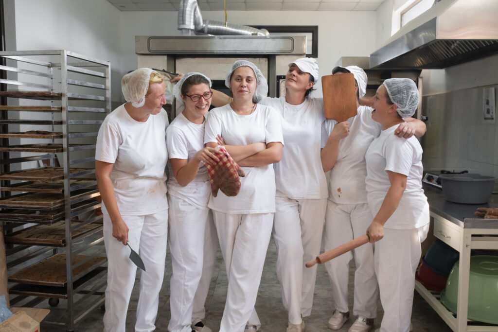Six women, all bakers, stand together in a commercial kitchen posing and laughing