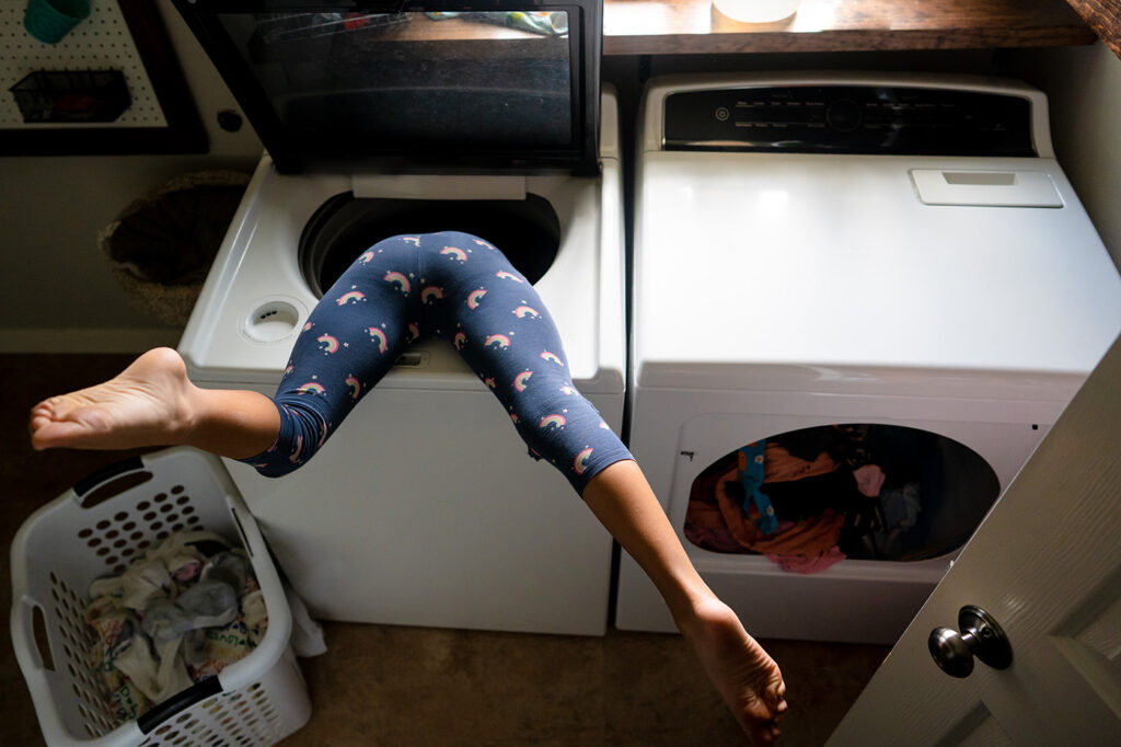 Laundry room scene with child helping with chores. Her legs kick in the air as she is stuck upside down in washing machine.