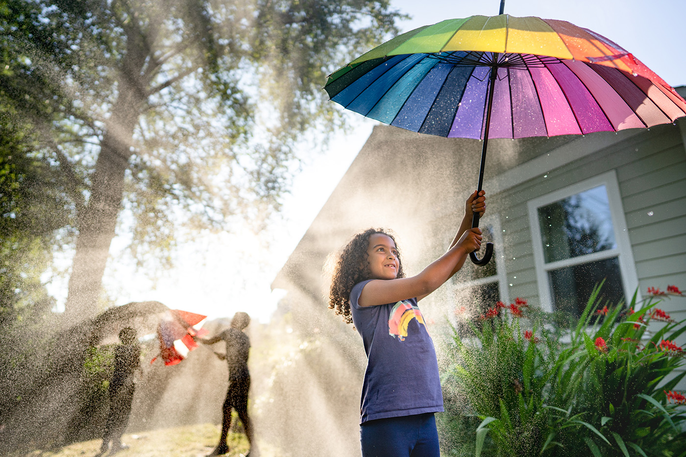 Curly haired girl holds rainbow umbrella aloft while two figures with umbrellas stand in the background. Air is full of water spray but sun is shining.