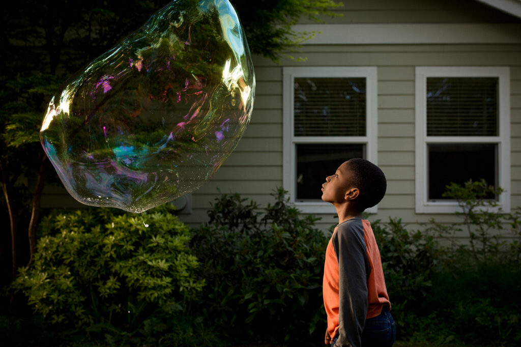 Boy looks in amazement at huge bubble with rainbow colors playing across its surface. He stands in front o fa house with lush foliage.