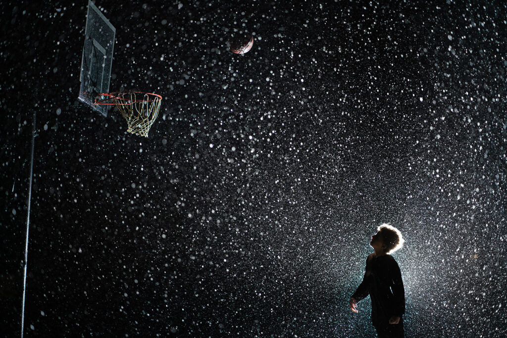 Silhouette of child shooting hoops in the rain. He is backlit with dramatic light, with raindrops filling the screen.