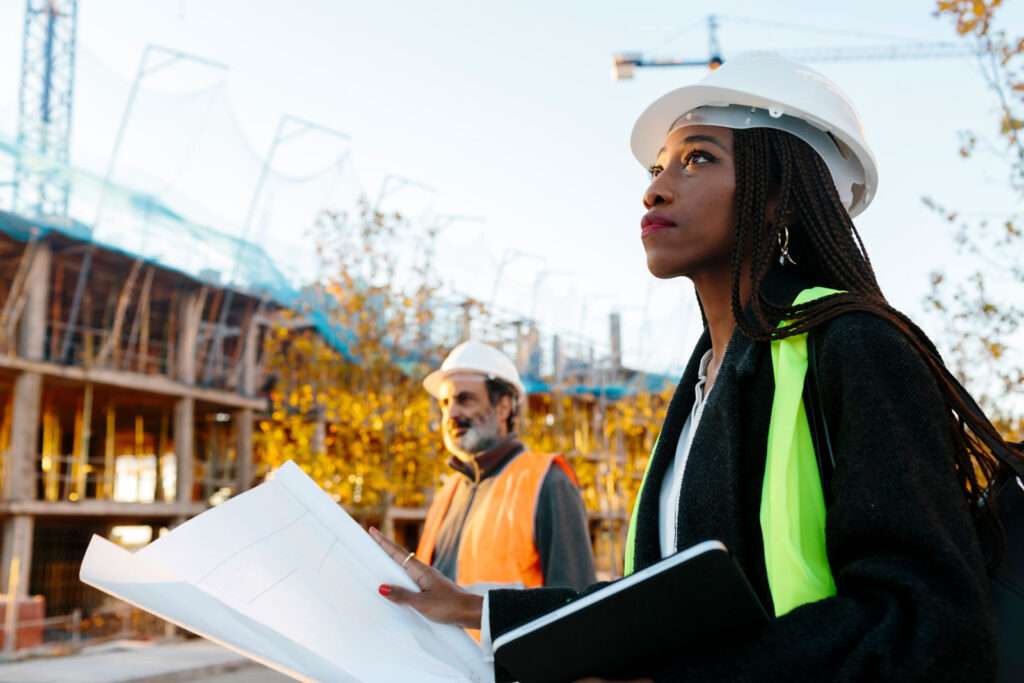 Professional architect woman supervising construction area. A worker from the construction is in the background.. They wear safety protective helmet and vest. It's sunset.