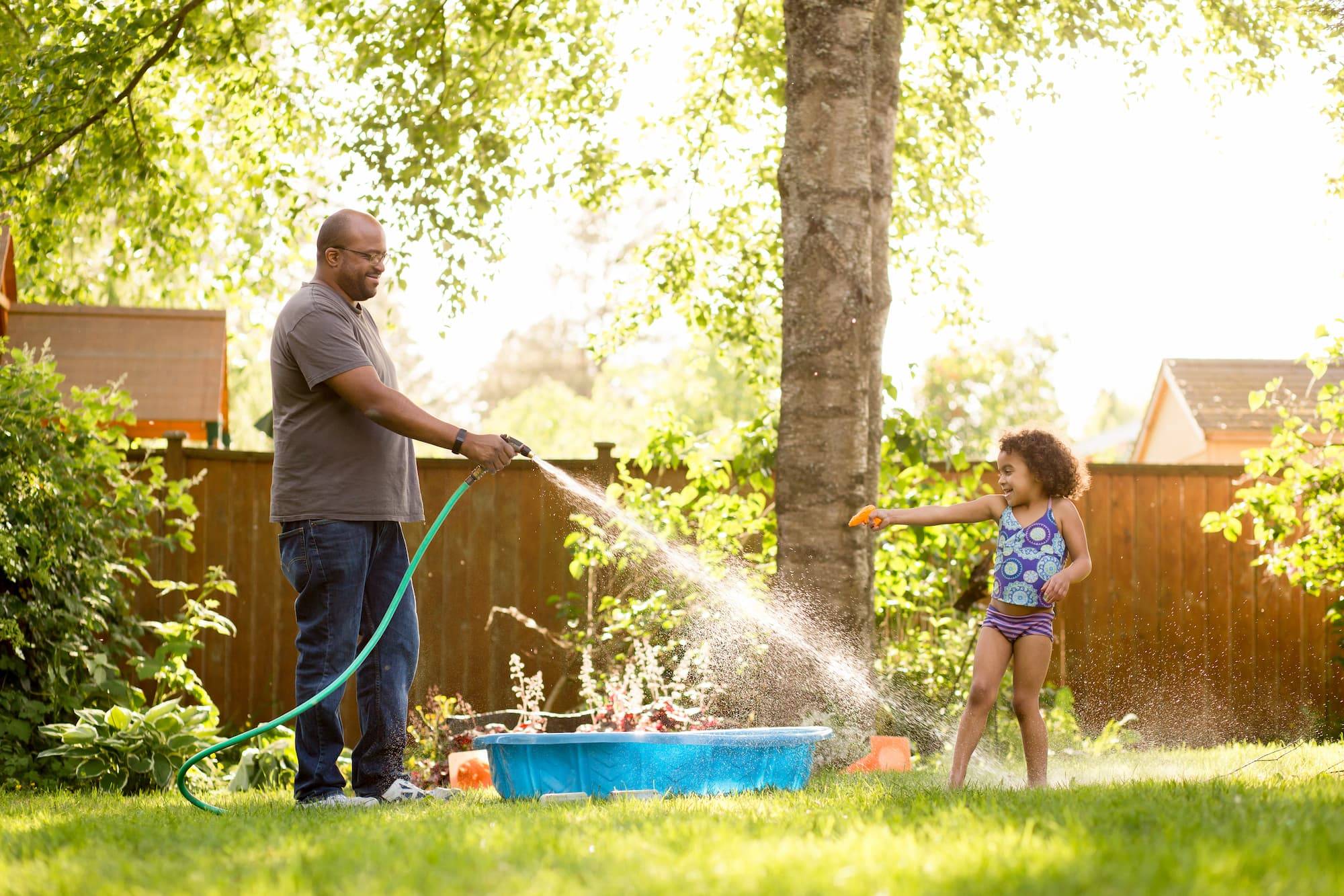 Black father and daughter have water fight in backyard. Father has a hose and daughter carries a small water pistol. Joyful summertime playful family scene.