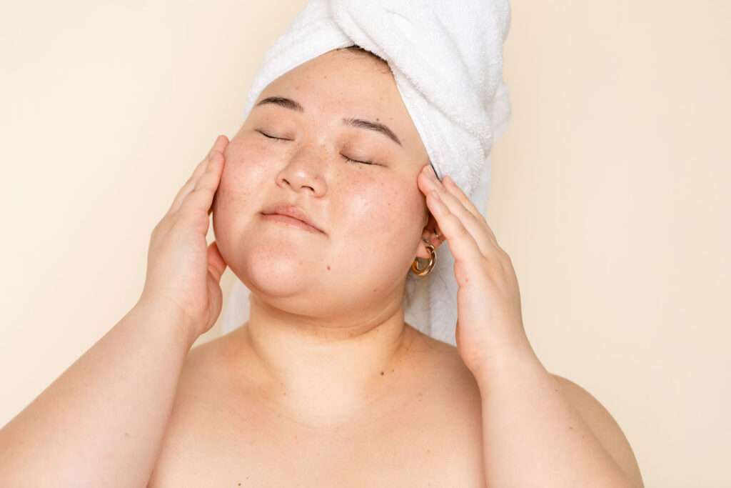 Asian woman with eyes closed doing her skincare routine on a fresh face after a shower.