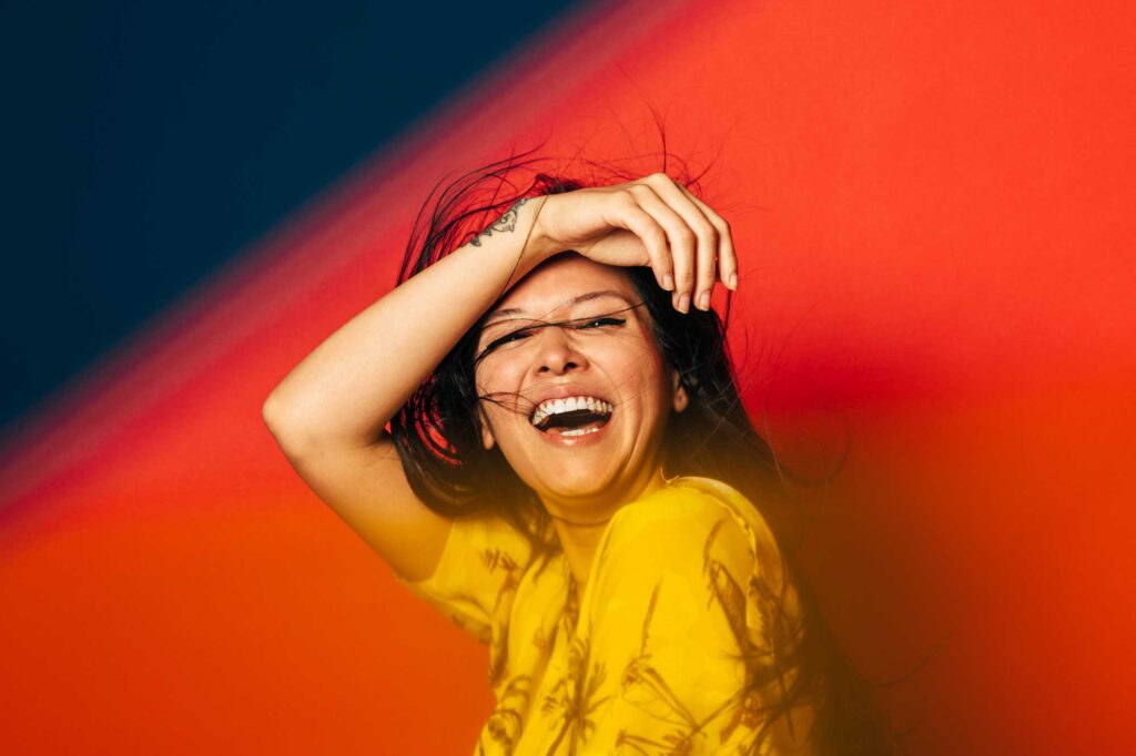 Happy young asian woman laughing over red background. She is wearing a yellow tropical shirt. She is covering her face with her arm.