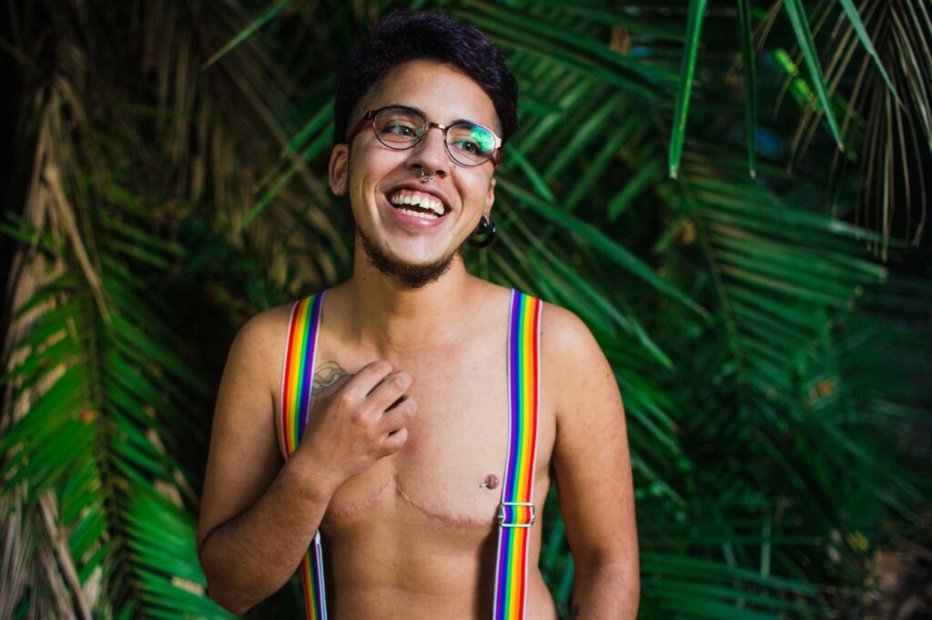 Young transgender man portrait in a nature environment wth tree leafs, green and dried. He is standing among the leaves wearing jeans and bare chest with lgbt rainbow flag colors suspenders.