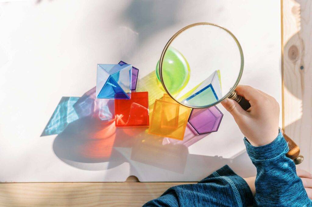 table set up for Montessori math learning with 3-d geometric shapes in a home learning environment for preschool homeschool- kid holds up magnifying glass