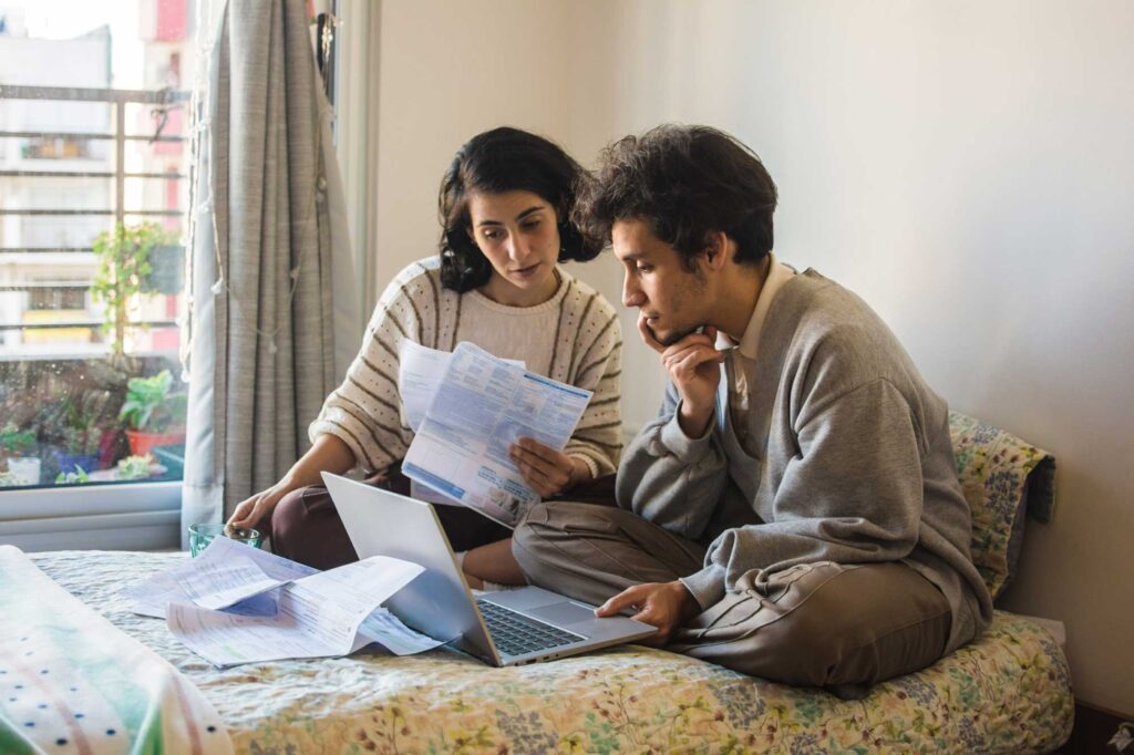 Young latino couple sitting in a mattress on the floor working on their personal and home finance. They are drinking coffee in a sunshine filled afternoon.