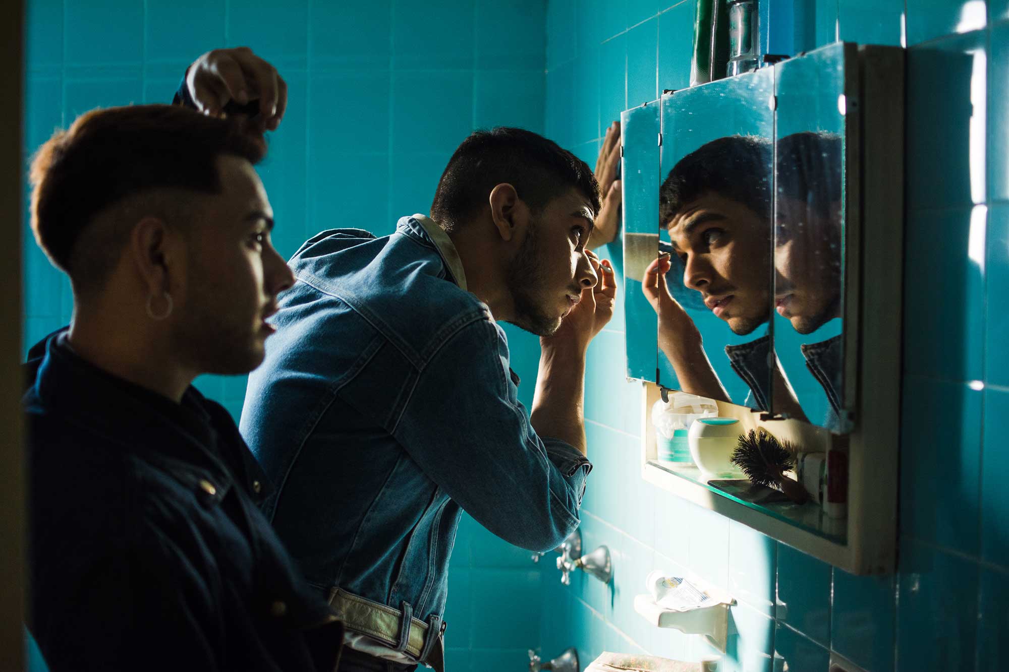Gay latino couple in the bathroom grooming and getting ready before going out. They are in denim outfits in a blue tiled bathroom with a sink and mirror.