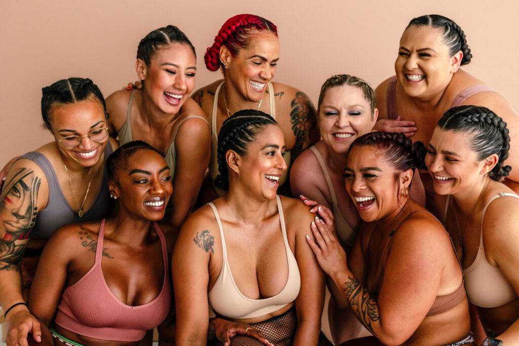 Close up of laughing female friends with tattoos and braided hair wearing only undergarments in front of neutral studio backdrop.