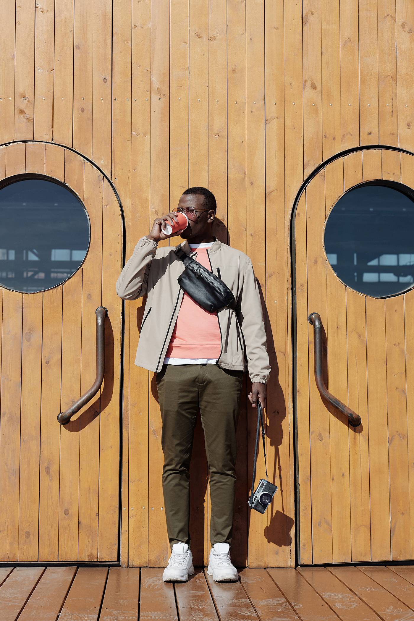 Full shot of young man drinking coffee standing against wooden building between two doors