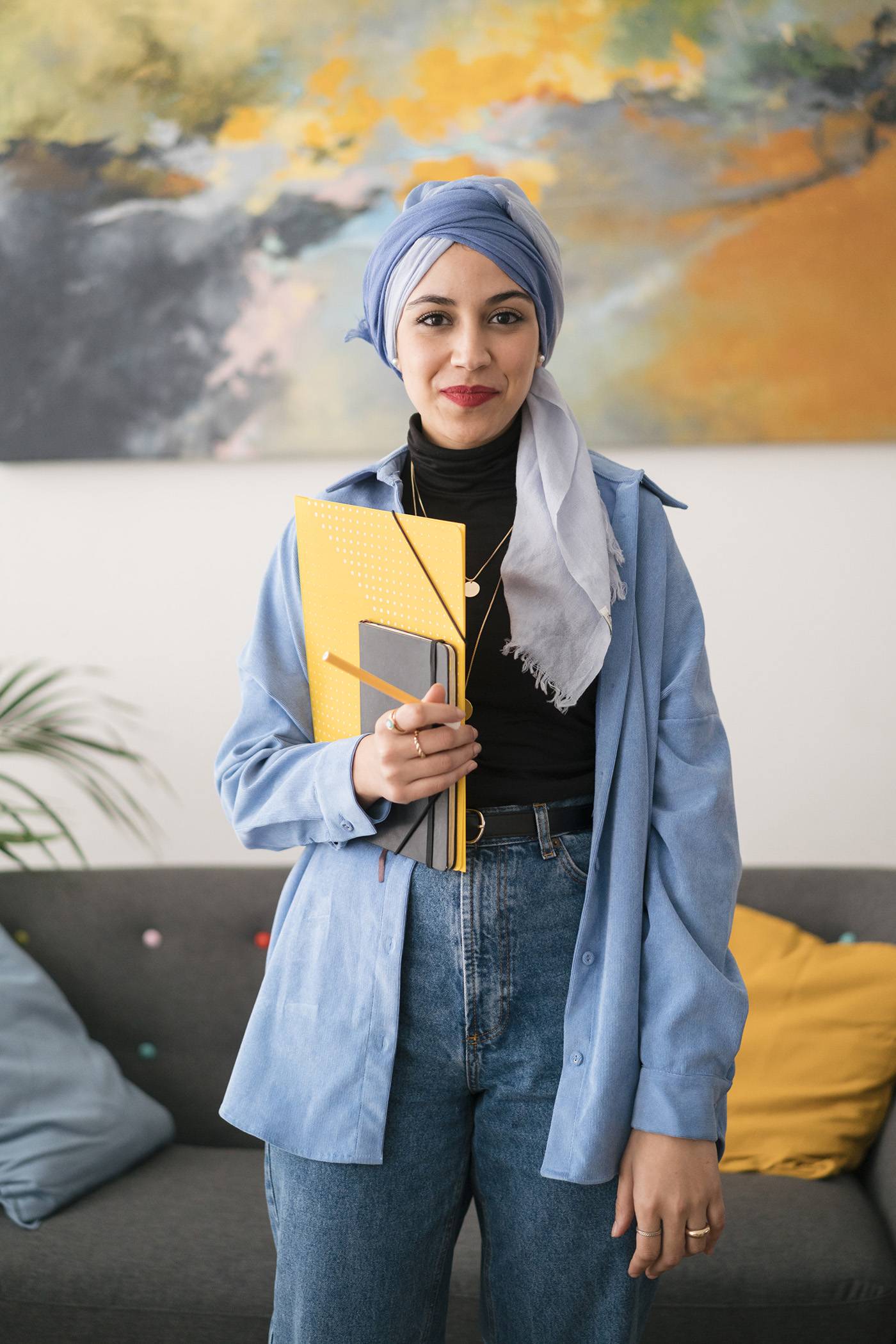 Positive female office worker in casual wear and headscarf holding papers and diary and looking at camera while standing in stylish room