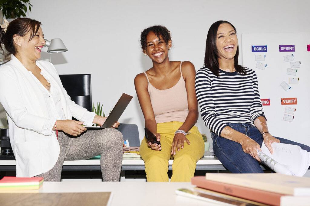 Diverse women in casual clothes with gadgets and documents sitting at table and laughing at funny joke during work on project in office.