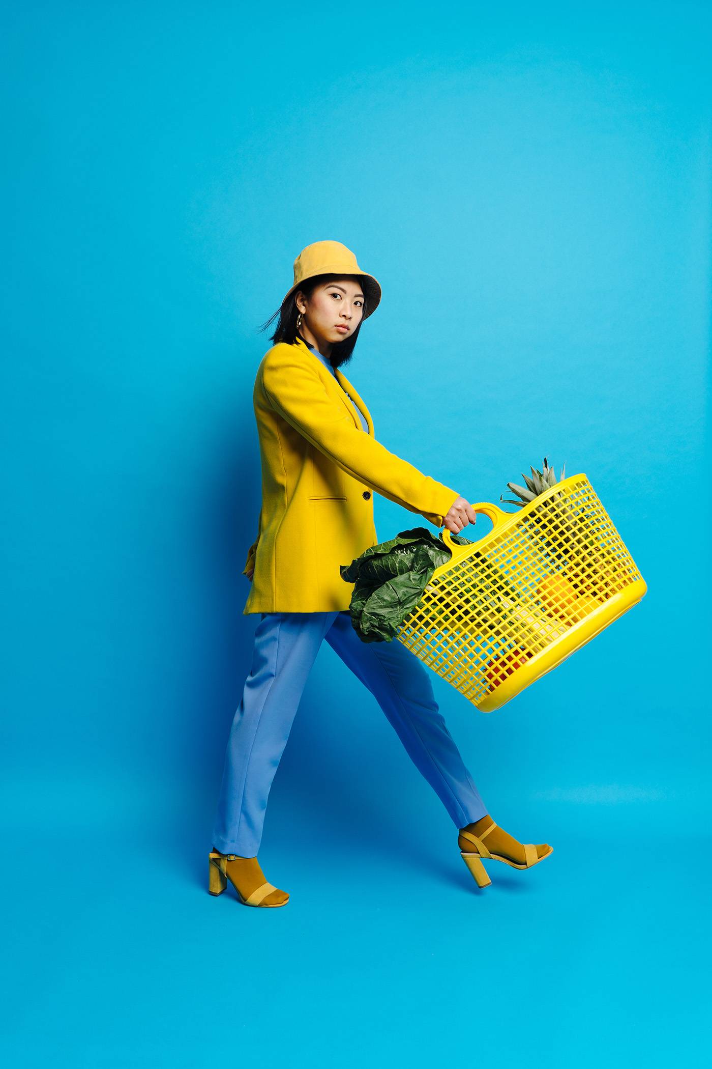Side view of elegant Asian teen in hat holding shopping basket in stretched out hand on blue background