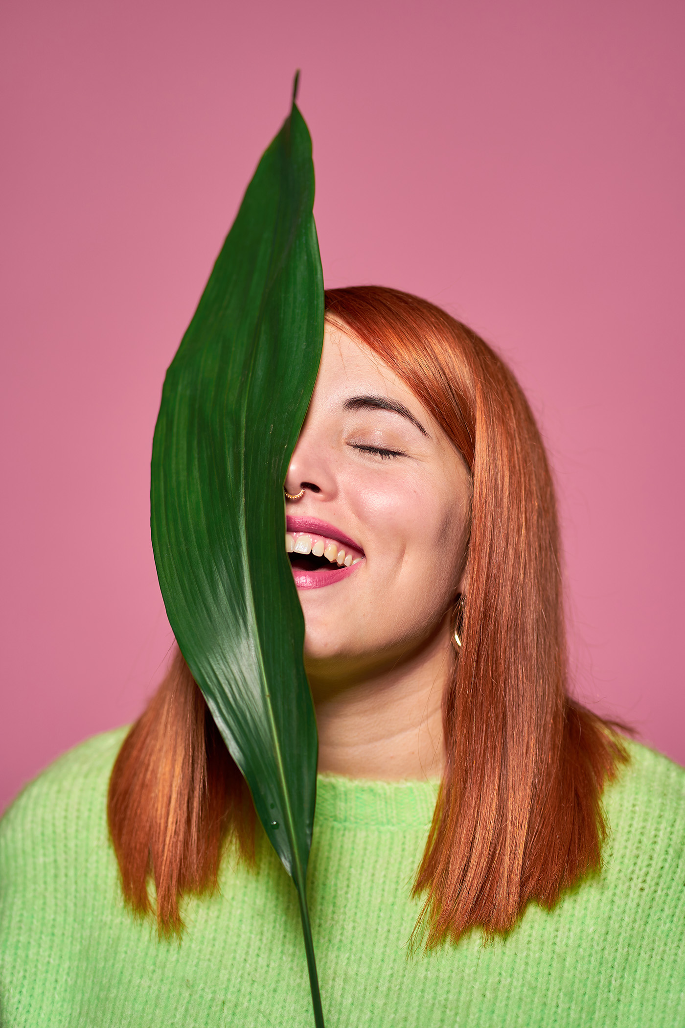 Curvy model with red hair keeping green leaf near face, laughing and eyes closed against pink background