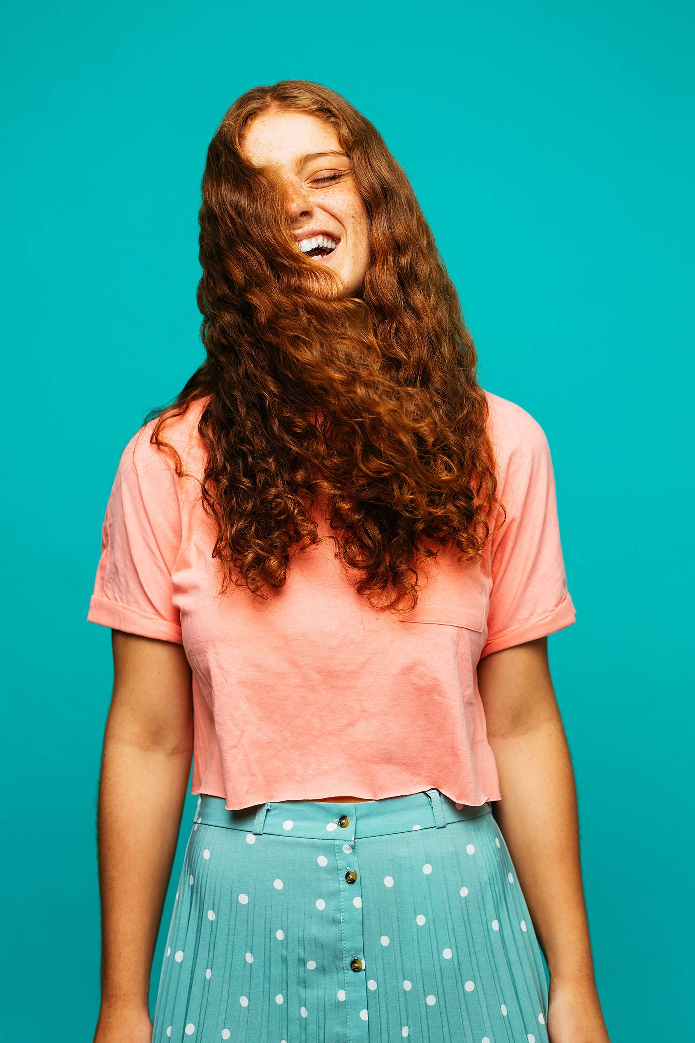 Joyful ginger teen girl with long curly hair covering face wearing pink blouse and blue polka dot plisse skirt laughing out loud while standing against turquoise background