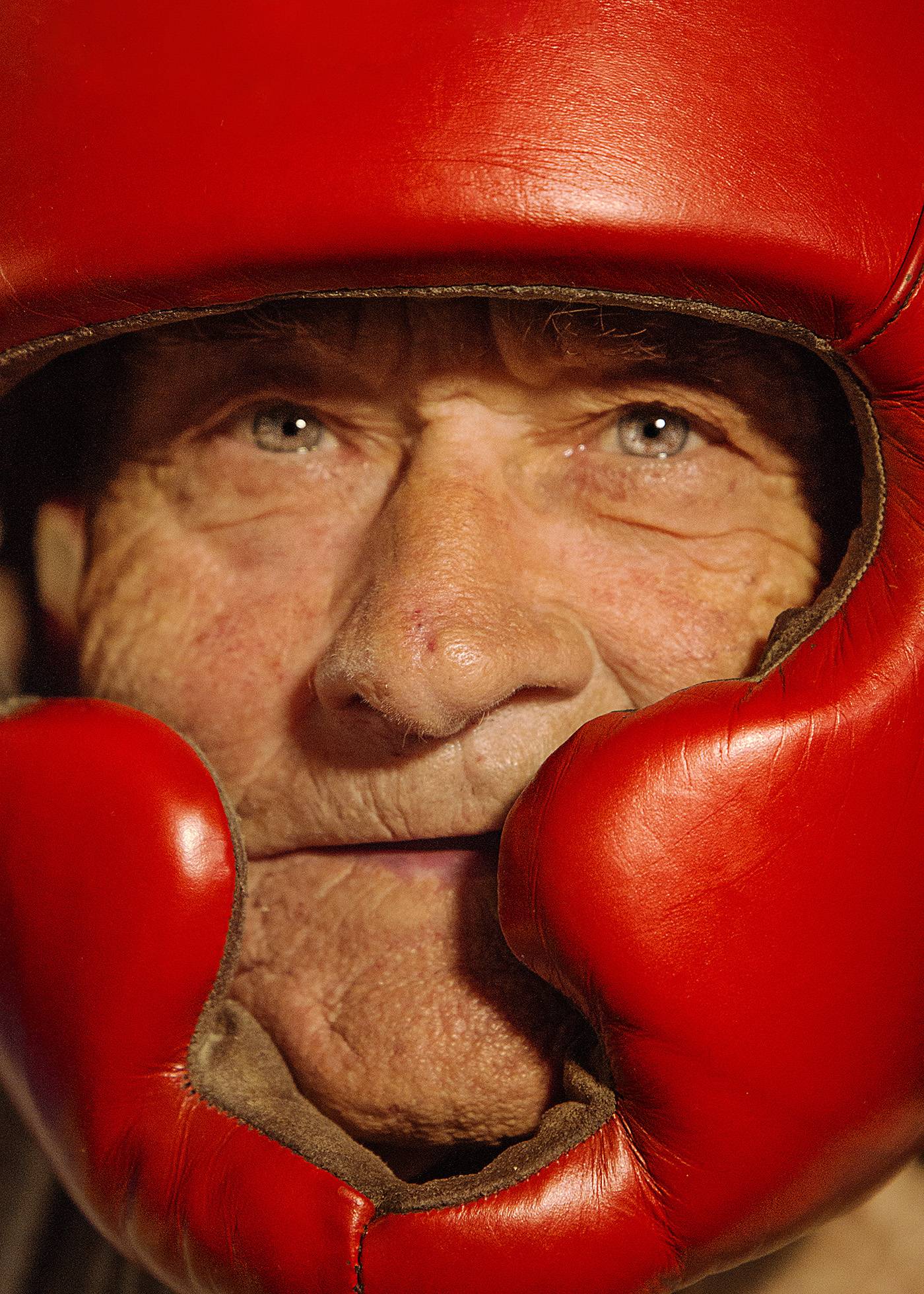 A portrait of an old man's face boxing. The man has a kind face and is looking into the camera with very fierce eyes. The old man is wearing red headgear and is ready to fight his opponent.